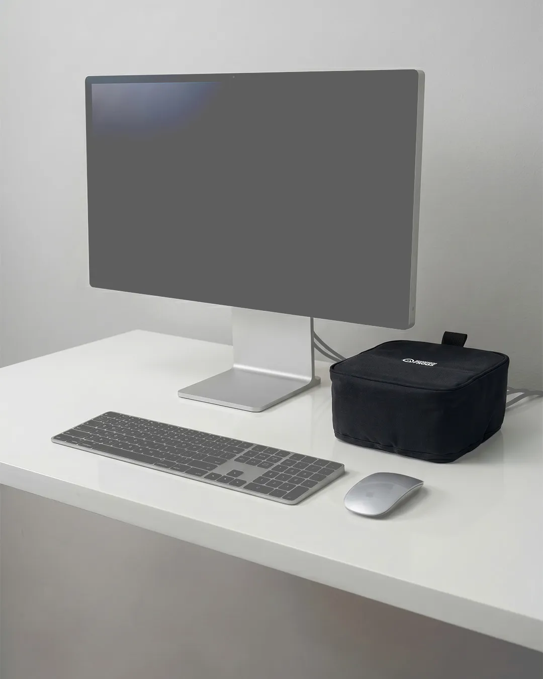 Minimalist modern home office desk setup with a silver desktop monitor, magic keyboard, mouse, and a covered Mac Studio in a black fabric sleeve.